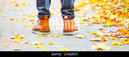 legs of a man in brown boots on the sidewalk strewn with fallen leaves. The concept of turnover of the seasons of the year. Weather background Stock Photo
