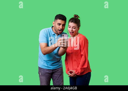 Friends reading shocking news! Portrait of amazed attractive young couple in casual wear reading unexpected message with open mouth, using mobile phon Stock Photo