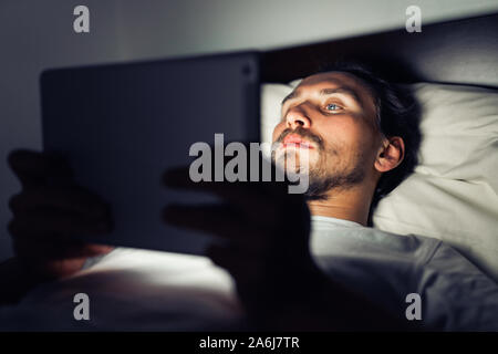 Young handsome and tired man with a beard cannot sleep and is watching something on his tablet at night. Stock Photo