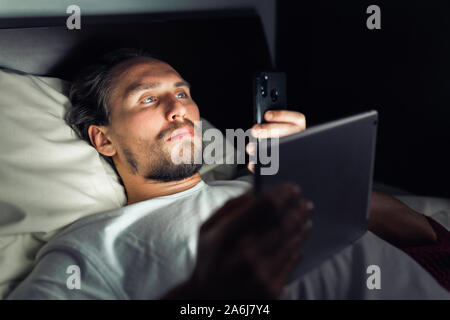 Young bearded man lying in bed while looking at his phone and tablet. Stock Photo