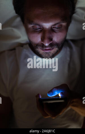 Young handsome and tired man with a beard cannot sleep and is watching something on his phone at night. Stock Photo