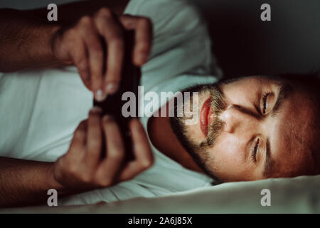 Young happy man cannot sleep and is watching something on his phone at night. White light from the phone is all over his face. Stock Photo