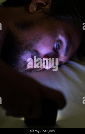 Bearded young handsome and tired man cannot sleep and is watching something on his phone at night. Blue light from the phone is all over his face. Stock Photo