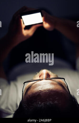 Bearded young man in glasses is lying in his bed at night while watching something on his mobile phone. Stock Photo