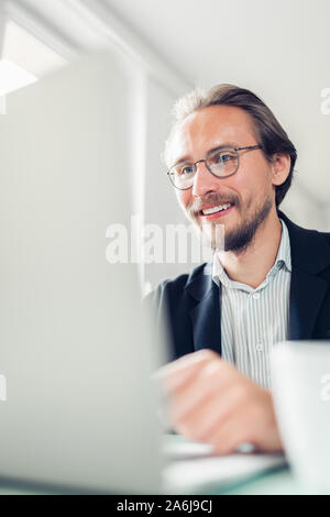 Photo of a handsome focused and smiling young man sitting by the desk working at the computer. Stock Photo