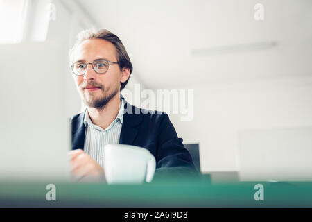 Photo of a handsome focused young man sitting by the desk and working at the computer. Stock Photo