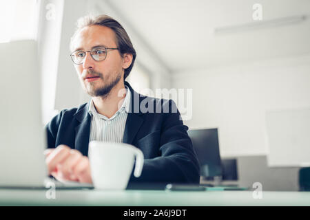 Photo of a handsome focused young man sitting by the desk and working at the computer. Coffee cup visible in the foreground.