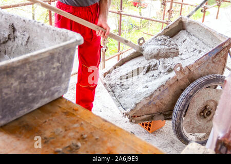 Construction worker is using shovel to unloading cement, mortar, from wheelbarrow on construction site. Stock Photo