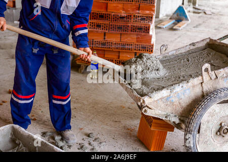 Construction worker is using shovel to unloading cement, mortar, from wheelbarrow on construction site. Stock Photo