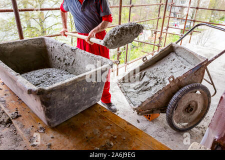 Construction worker is using shovel to unloading cement, mortar, from wheelbarrow on construction site. Stock Photo