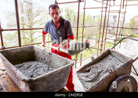 Construction worker is using shovel to unloading cement, mortar, from wheelbarrow on construction site. Stock Photo