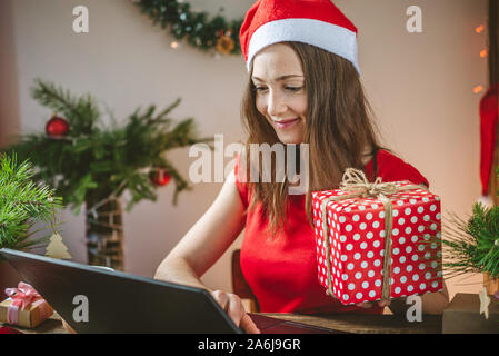 A beautiful woman in a red dress is holding a gift box and ordering online purchases on her laptop. Online shopping for Christmas holidays. Stock Photo