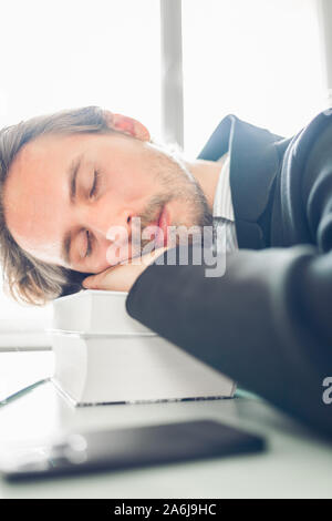 Handsome young man sleeping on pile of books at work desk. Phone and a laptop on a desk next to him. Stock Photo