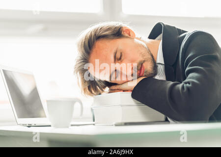 Handsome young man sleeping on pile of books at work desk. Coffee, phone and a laptop on a desk next to him. Stock Photo