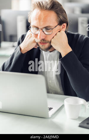 Tired and bored young handsome man working at the computer. Coffee cup visible next to him. Stock Photo