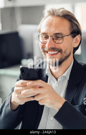 Young handsome man in glasses is interacting with his mobile phone while being at work. Happiness and content at work and being always reachable conce Stock Photo