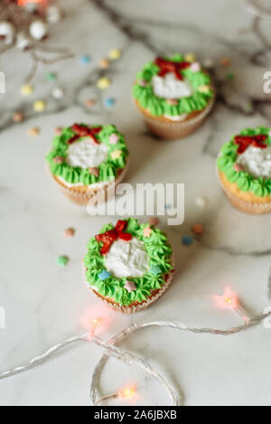 Christmas cupcakes with green Christmas wreath of cream on marble background with garland. Christmas sweets decorated with mastic. Stock Photo