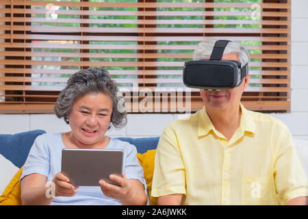 Asian senior couple play virtual realtiy glasses headset and tablet watching vr video and  have fun together on sofa  in living room at home.senior wi Stock Photo