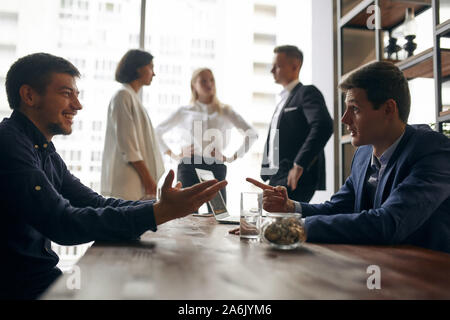 cheerful two men in stylish formal clothes having a cunning plan, handsome man pointing to his colleague, teaching him to run business Stock Photo