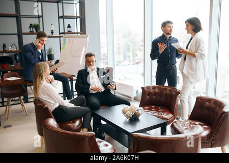 young creative people are discussing work. Concept of a busy office life.handsoem serious guy sitting on the armchair holding smart phone, blonde woma Stock Photo