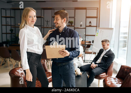 smiling blonde woman in white shirt and black trousers putting her hand on her colleague shoulder while he is looking at her and laughing. Stock Photo