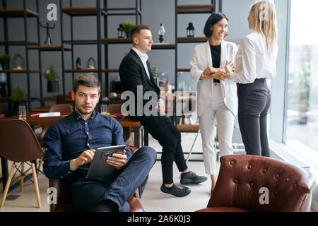 handsome guy sitting on the armchair with tablet, looking at the camera while his three colleagues having fun in the background of the photo. Stock Photo