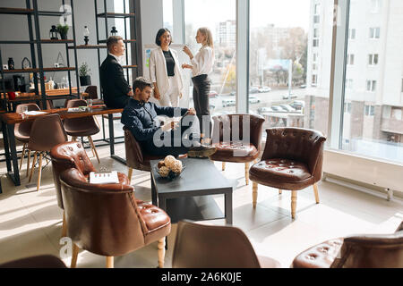 serious handsome man sitting with crossed arms working with tablet while his colleagues having a conversation behind him. copy space. panorama window. Stock Photo
