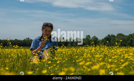 Teen girl holding flowers and smiling while sitting in large field with yellow flowers and trees in the background Stock Photo
