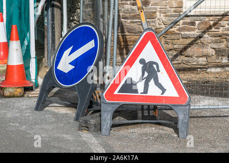 Man at Work pictogram roadworks sign in town of Lostwithiel, Cornwall, with blue Left arrow pointing traffic direction sign. Digging up road. Stock Photo