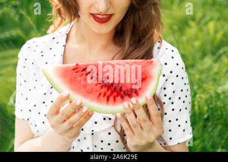 Smiling girl holds slice of watermelon on green grass background. Smiling young woman is holding slice of watermelon close up. Stock Photo