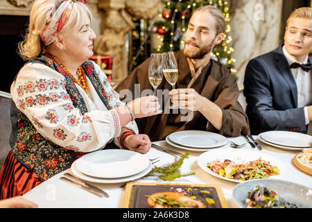 Elegantly dressed group of people having a festive dinner at a well-served table near the christmas tree, celebrating New Year holiday at the luxury restaurant Stock Photo