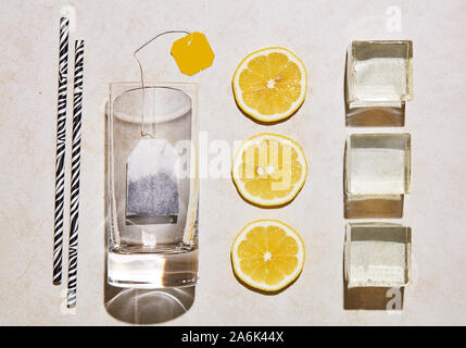 Ice tea ingredients. Glass, straw, lemon, ice cubes and teabag under hars summer sunlight. Top view. Stock Photo