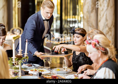 Elegantly dressed group of people having a festive dinner during New Year Eve at the luxury restaurant, waiter serving dishes Stock Photo