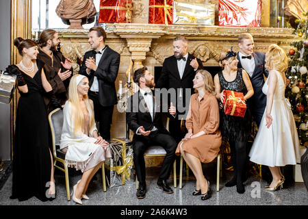 Staged group photo of an elegant well-dressed people near the beautifully decorated fireplace with christmas tree and presents during a New Year celebration Stock Photo
