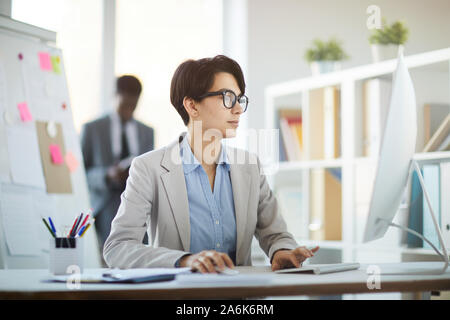 Side view portrait of successful young businesswoman using PC while sitting at desk in office and enjoying work, copy space Stock Photo