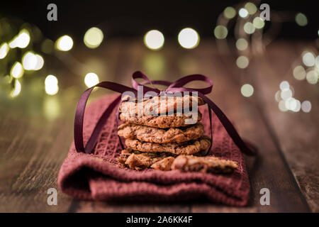 A stack of delicious chocolate chip cookies tied with a purple ribbon, on a matching napkin. On a wooden table with string lights in the background Stock Photo