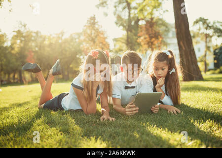 three smiling school-age friends are lying on the grass and looking at the tablet Stock Photo