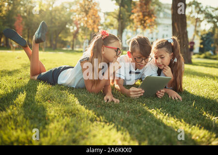 three smiling school-age friends are lying on the grass and looking at the tablet Stock Photo