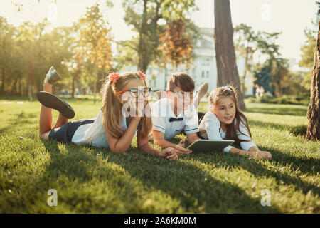 Three smiling school-age friends are lying on the grass and looking at the camera. In the hands of children a tablet Stock Photo