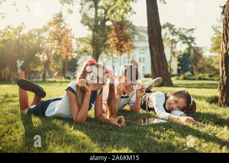 Three smiling school-age friends are lying on the grass and looking at the camera. In the hands of children a tablet Stock Photo