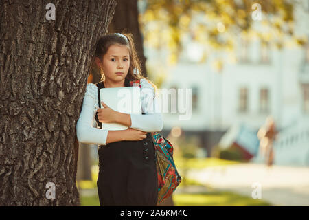 A beautiful girl of school age is standing by a tree and holding a tablet. Schoolgirl dressed in a sundress Stock Photo
