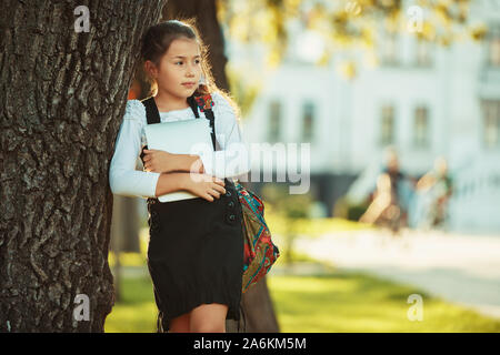 A beautiful girl of school age is standing by a tree and holding a tablet. Schoolgirl dressed in a sundress Stock Photo