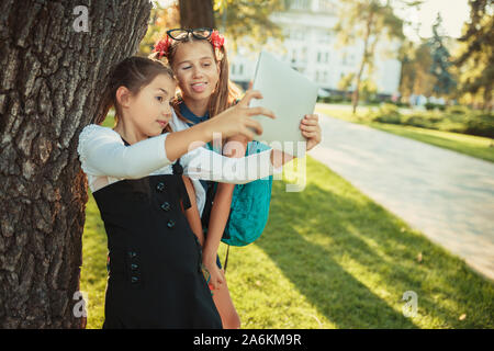 Two charming school-age girls are standing by the tree next to the school and playing on the tablet. schoolgirls take a selfie Stock Photo