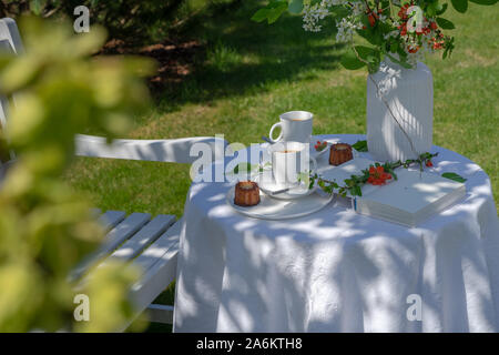 Enjoying coffee time on a sunny spring day. Served table with coffee, canele and flowers in the garden. There is a white wooden bench next to it Stock Photo