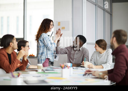 Portrait of creative business team giving high five while planning project during meeting in office, focus on smiling young woman standing by table Stock Photo