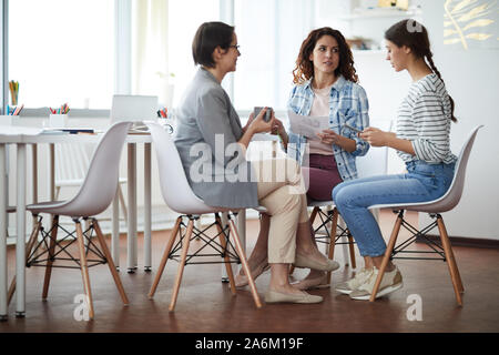 Full length view at three contemporary women discussing photographs sitting at table against window, copy space Stock Photo