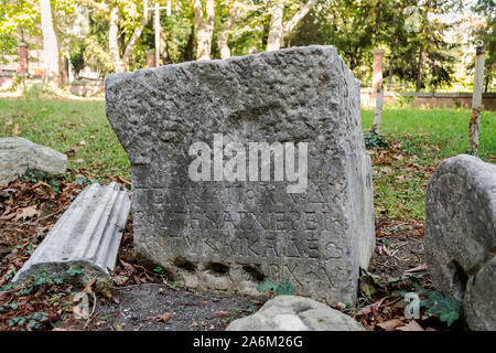 Fragment of the ancient carved stone block with an inscription in Greek. Antique artifact outdoors. On the green grass. Stock Photo