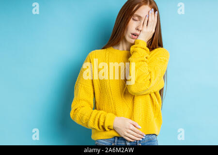 Shot of of tired overworked caucasian redhead female covering face with palm, closing eyes, having sleepy expression, dressed in yellow jumper isolate Stock Photo