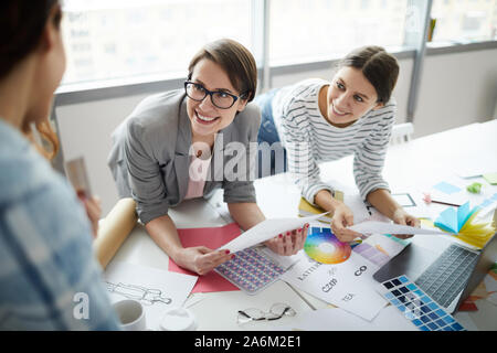High angle portrait of two smiling businesswomen discussing creative project leaning over table with photographs and color swatches, copy space Stock Photo