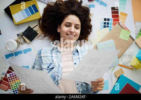 Above view portrait of creative young woman smiling happily while lying on floor surrounded by designer supplies, copy space Stock Photo